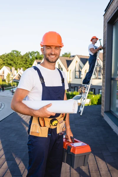 Selective focus of builder in tool belt holding blueprint and toolbox near colleague on roof — Stock Photo