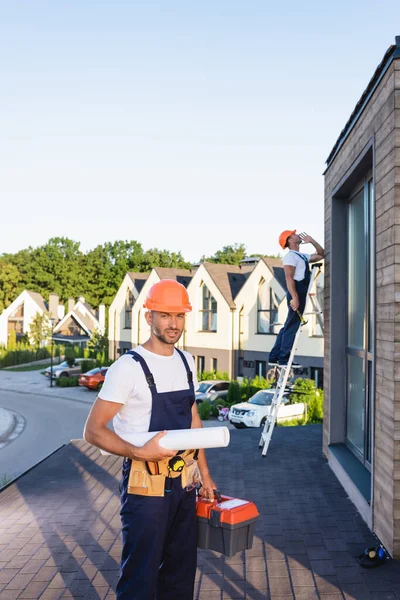 Selektiver Fokus des Bauherrn mit Werkzeugkasten und Bauplan auf dem Dach des Gebäudes — Stockfoto