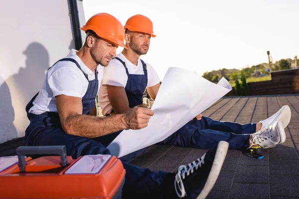 Selective focus of builders with blueprint sitting near toolbox while working on roof of house — Stock Photo