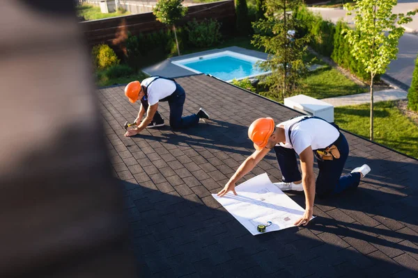 Selective focus of builders with blueprint and tools working on roof of building — Stock Photo