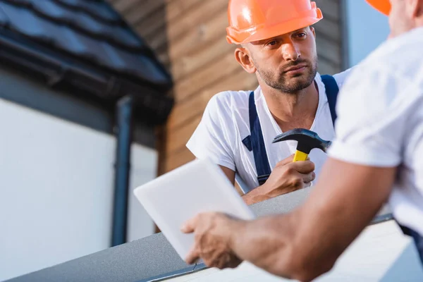 Selective focus of builder with hammer looking at colleague with digital tablet on roof of building — Stock Photo