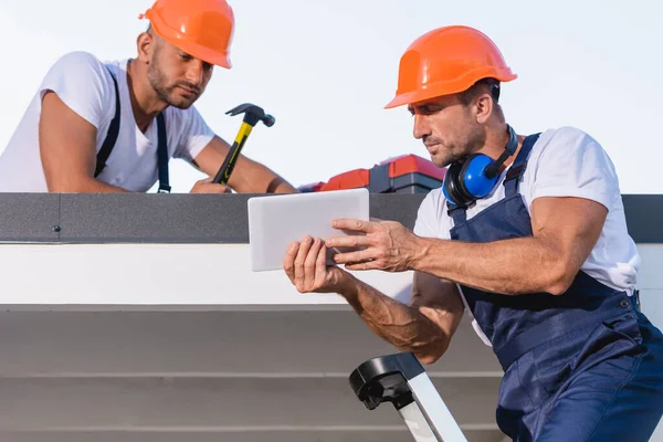 Reparadores em macacões e capacetes usando tablet digital no telhado da casa — Fotografia de Stock