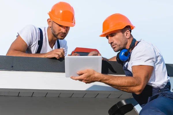 Selective focus of handymen in workwear using digital tablet while working on roof of building — Stock Photo