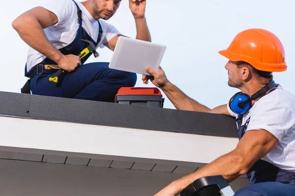 Selective focus of builder showing digital tablet to colleague with hammer on roof of building — Stock Photo