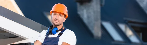 Panoramic shot of builder in hardhat looking at camera near roof of building — Stock Photo