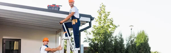 Horizontal crop of builders looking at camera while using ladder near building — Stock Photo