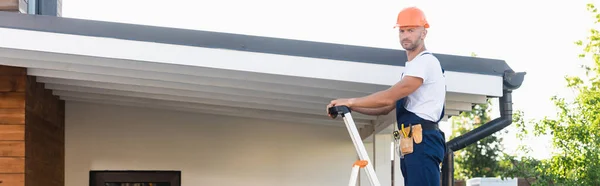 Панорамний знімок будівельника, який дивиться на камеру, стоячи на сходах біля даху будівлі — стокове фото