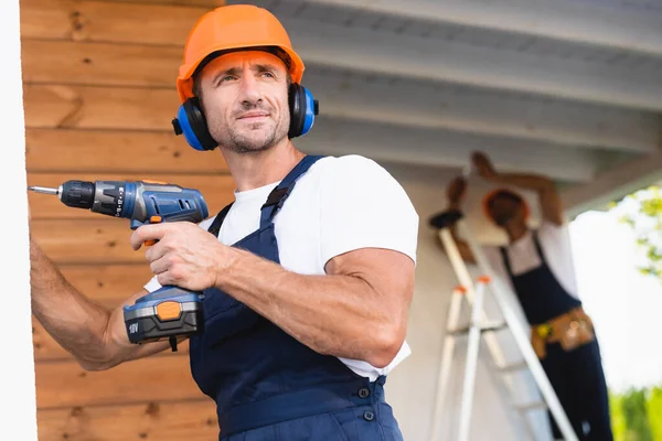 Selective focus of handyman in workwear and ear defenders holding electric screwdriver near facade of building — Stock Photo