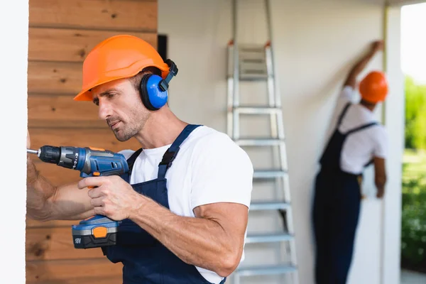 Selective focus of handyman in hardhat and uniform using electric screwdriver on facade of building — Stock Photo
