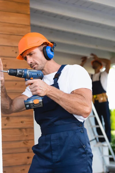 Selective focus of manual worker in helmet and ear defenders holding electric screwdriver near facade of house — Stock Photo