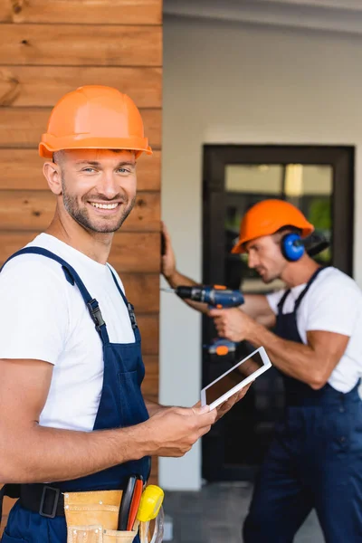 Selective focus of builder holding digital tablet near colleague working with electric screwdriver on facade of building — Stock Photo