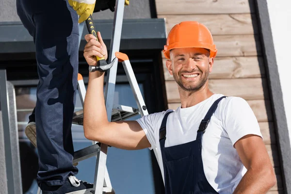 Selective focus of builder looking at camera while giving hammer to colleague on ladder near house — Stock Photo