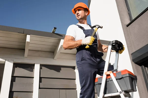 Bauarbeiter in Overalls und Handschuhen hält Hammer neben Werkzeugkiste auf Leiter und Gebäude — Stockfoto