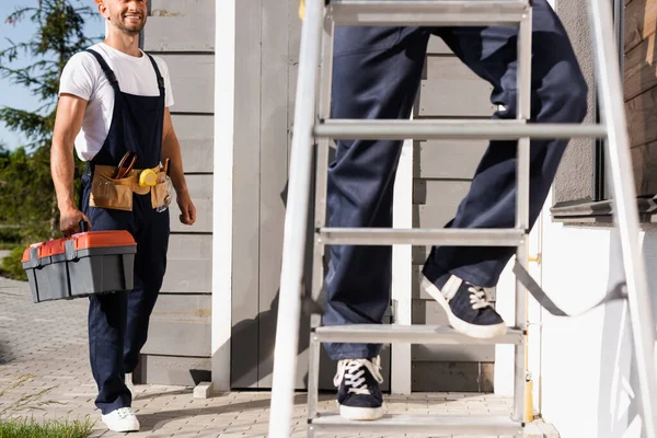 Enfoque selectivo de constructor sosteniendo caja de herramientas cerca de colega en escalera cerca de casa - foto de stock