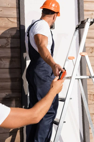 Selective focus of builder giving pliers to colleague on ladder near house — Stock Photo