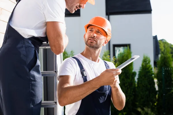 Selektiver Fokus des Bauunternehmers in Overalls mit digitalem Tablet in der Nähe des Kollegen auf Leiter und Haus — Stockfoto