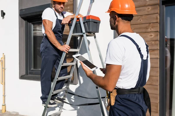 Enfoque selectivo del manitas con tableta digital de pie junto a su colega en uniforme en escalera y casa - foto de stock