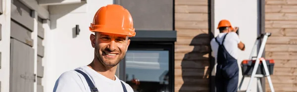 Panoramic shot of builder looking at camera while colleague working near building at background — Stock Photo