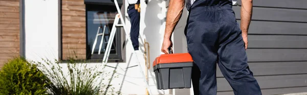 Panoramic crop of builder in uniform holding toolbox near house outdoors — Stock Photo
