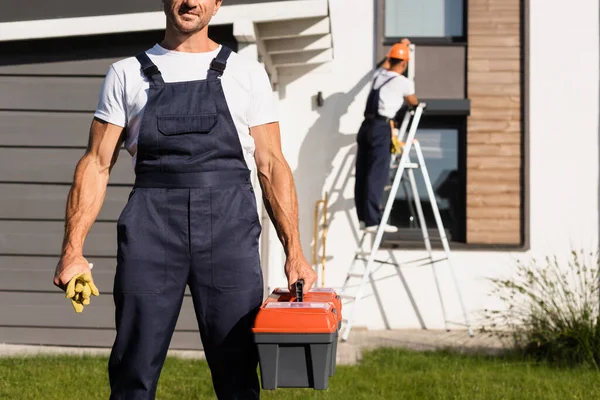 Focus selettivo del tuttofare in tuta con guanti e cassetta degli attrezzi sul prato vicino alla casa — Foto stock