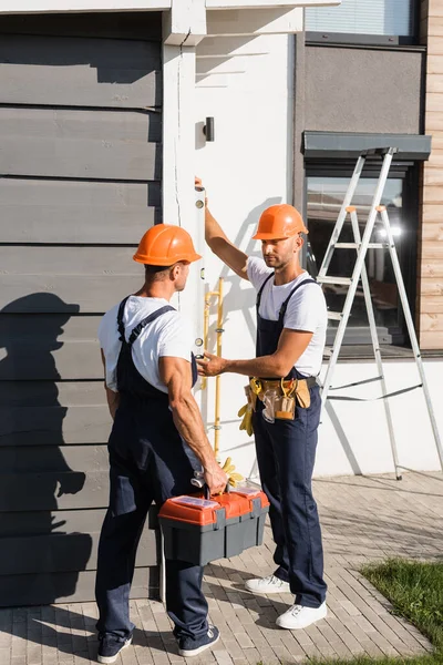 Costruttore in cassetta portautensili uniforme vicino al collega con livella e edificio — Foto stock