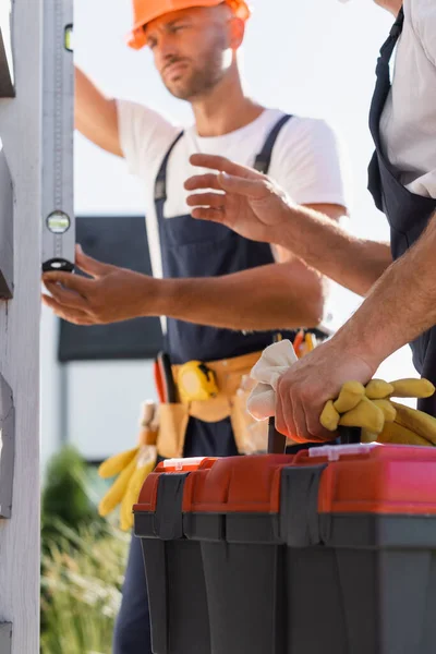 Selective focus of workman holding gloves and toolbox while colleague using spirit level near house — Stock Photo