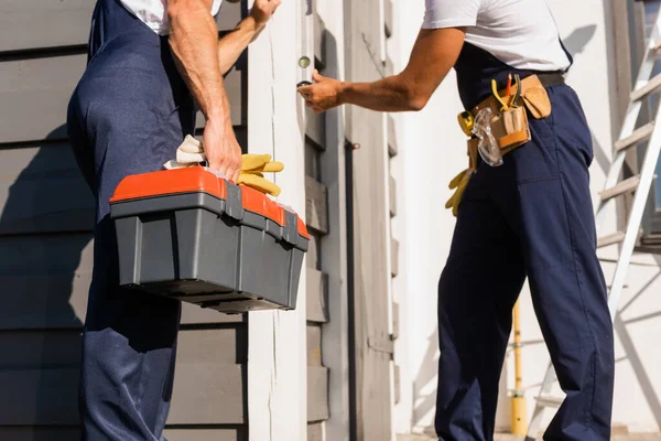 Cropped view of builder holding toolbox and gloves near colleague with spirit level and building — Stock Photo