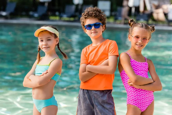 Ragazzo in occhiali da sole e ragazze in costume da bagno in posa con le braccia incrociate vicino piscina — Foto stock