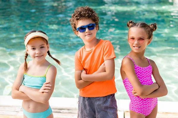 Девушки в купальниках и кудрявый мальчик в солнцезащитных очках, стоящие со скрещенными руками возле бассейна — стоковое фото
