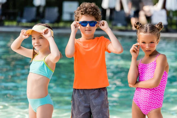 Menino encaracolado em t-shirt e meninas em maiôs tocando óculos de sol perto da piscina — Fotografia de Stock