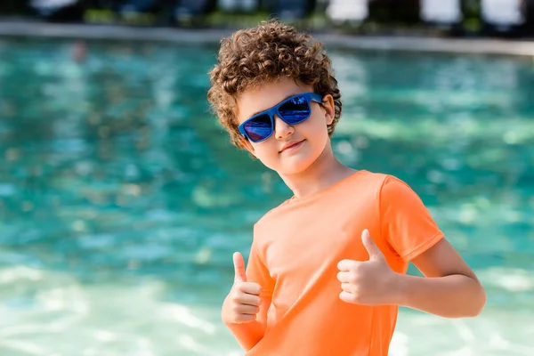 Curly boy in blue sunglasses and orange t-shirt showing thumbs up near pool — Stock Photo