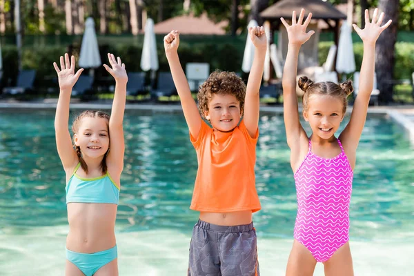 Девушки в купальниках и мальчик в футболке, стоящие с руками в воздухе возле бассейна — стоковое фото