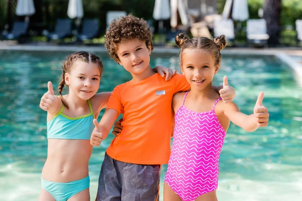 Menino encaracolado em t-shirt e meninas em maiôs mostrando polegares perto da piscina — Fotografia de Stock