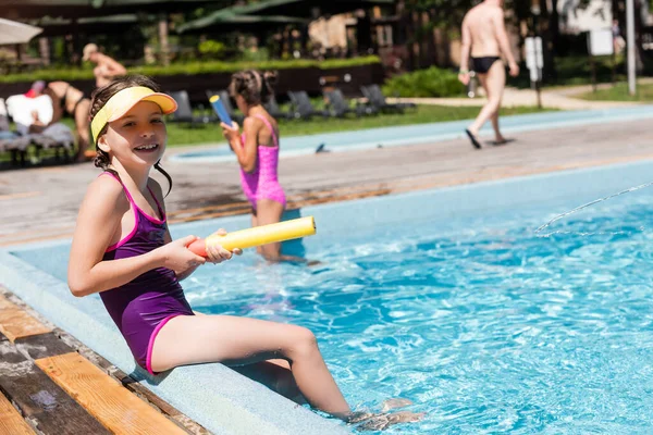 Selective focus of girl in swimsuit holding water gun and looking at camera while sitting at poolside — Stock Photo