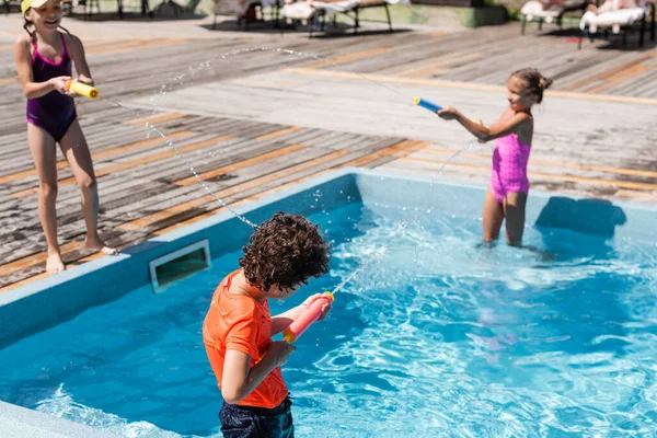 Selective focus of boy and two girls fighting with water guns near pool — Stock Photo
