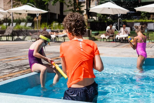 Visão traseira do menino se divertindo com amigos enquanto luta com armas de água perto da piscina — Stock Photo