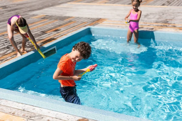 Вибірковий фокус хлопчика з закритими очима тримає водяний пістолет, розважаючись з дівчатами біля басейну — стокове фото