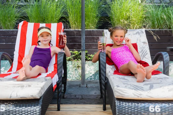 Girls in swimsuits looking at camera while sitting in sunbeds with refreshing cocktails — Stock Photo