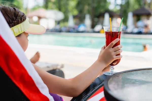 Selektiver Fokus des Mädchens mit Sonnenblendkappe, das Cocktailglas hält, während es sich im Liegestuhl ausruht — Stockfoto