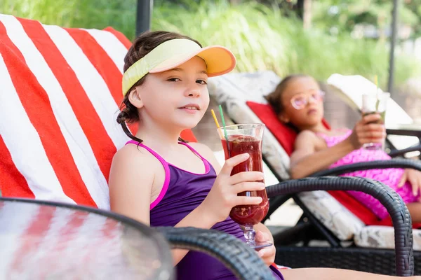 Selective focus of girl in sun visor cap sitting in sunbed near friend and holding glass of fresh cocktail — Stock Photo