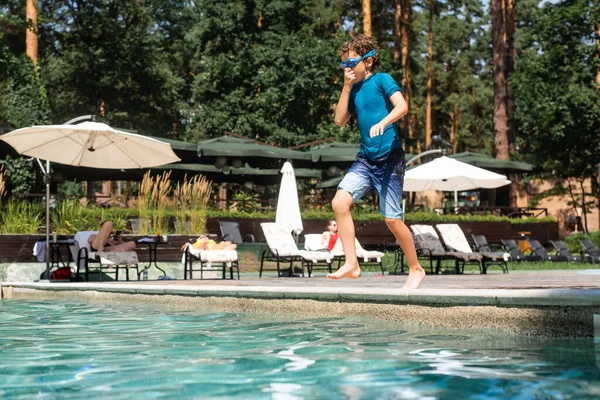 Niño en gafas de baño, camiseta y pantalones cortos tapando la nariz con la mano mientras salta a la piscina - foto de stock