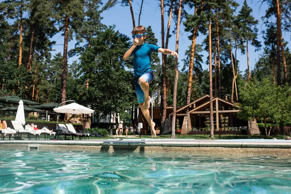 Kid in swim goggles, t-shirt and shorts plugging nose while jumping into swimming pool — Stock Photo