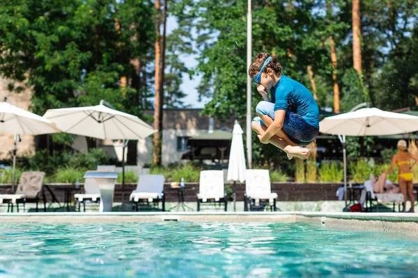 Vista lateral do menino em t-shirt e óculos de natação tapando o nariz enquanto salta para a piscina — Fotografia de Stock
