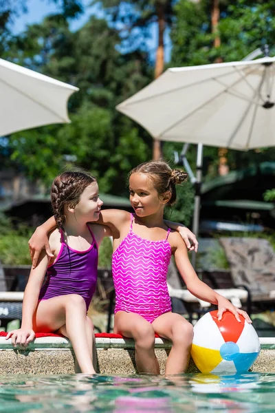 Meninas em maiôs sentados à beira da piscina com as pernas na água, abraçando e olhando um para o outro — Fotografia de Stock