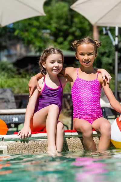 Niños en trajes de baño sentados junto a la piscina con las piernas en el agua, abrazando y mirando a la cámara - foto de stock