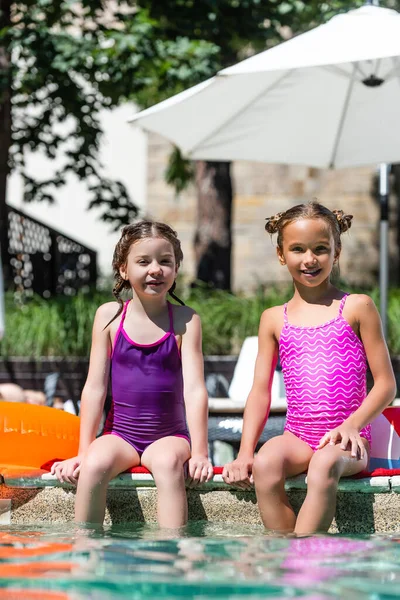 Amigos en trajes de baño sentados cerca de la piscina con piernas en el agua y mirando a la cámara - foto de stock