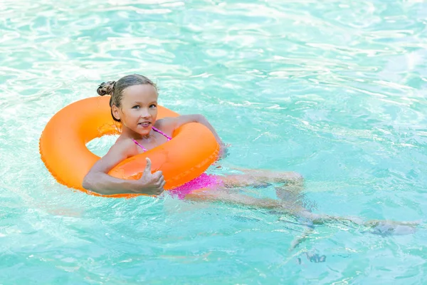 Chica complacida flotando en la piscina en el anillo inflable y mostrando el pulgar hacia arriba - foto de stock