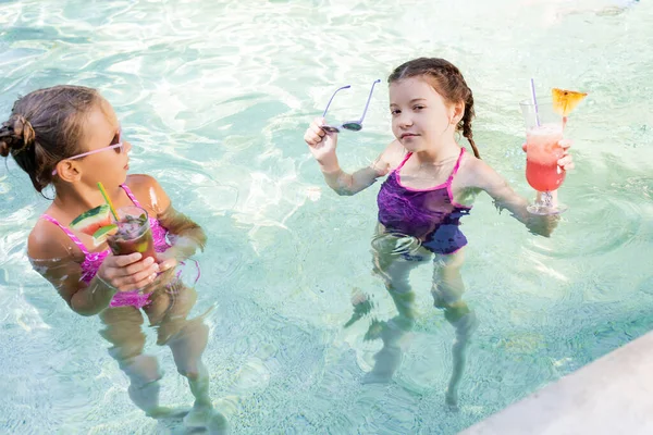 Girl looking at camera while holding sunglasses and fruit cocktail in pool near friend — Stock Photo