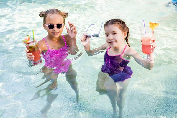 Kids in swimsuits holding fresh fruit cocktails in pool and looking at camera — Stock Photo