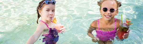 Fille en lunettes de soleil regardant la caméra tout en se tenant dans la piscine près d'un ami boire cocktail de fruits, concept horizontal — Photo de stock